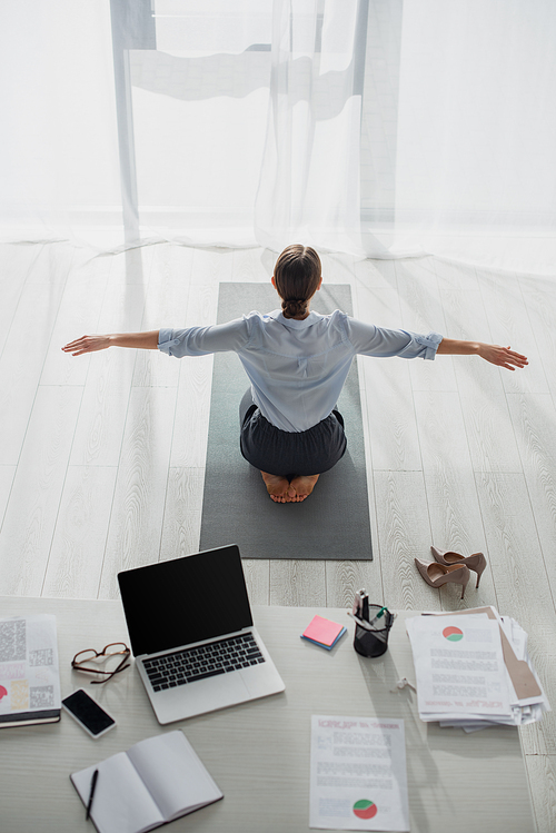 back view of businesswoman practicing yoga in lotus position on mat in office with laptop