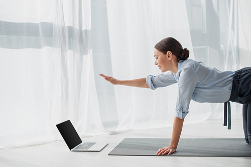 smiling businesswoman having online classes on laptop and practicing yoga in bird dog pose on mat in office