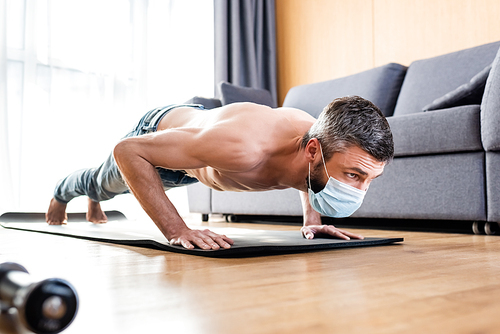 Selective focus of shirtless man in medical mask doing press ups on fitness mat in living room