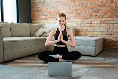 happy sportswoman with praying hands watching online yoga exercise on laptop in living room