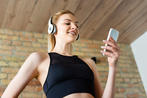 low angle view of cheerful sportswoman in wireless headphones using smartphone and standing with hand in pocket