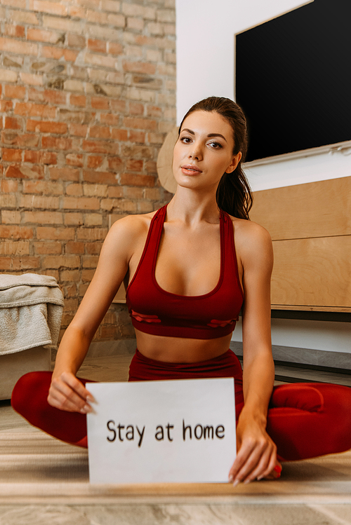 beautiful sportswoman holding stay at home sign while sitting on fitness mat at home during self isolation