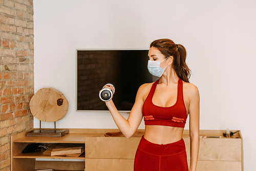 attractive woman in medical mask training with dumbbell at home during self isolation
