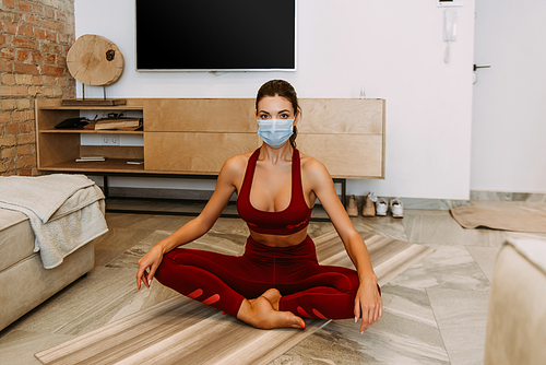 girl in medical mask sitting in lotus pose on yoga mat at home on quarantine