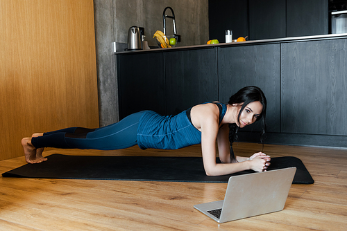 athletic woman in plank on fitness mat training online with laptop