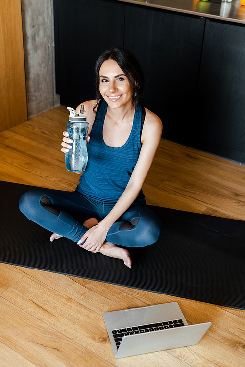 smiling sportive woman holding bottle of water while sitting on fitness mat with laptop