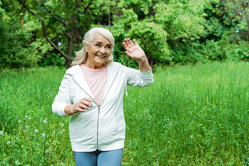 cheerful senior woman with grey hair waving hand in park