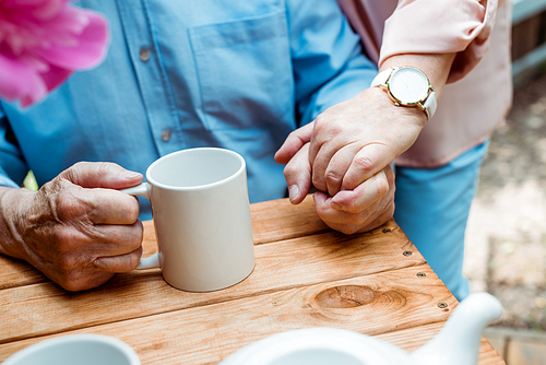 cropped view of senior man with cup of tea holding hands with retired wife