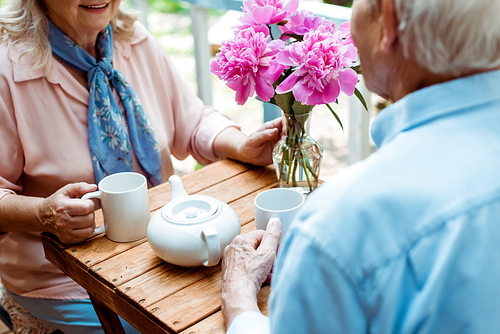 cropped view of senior couple holding cups while sitting near teapot