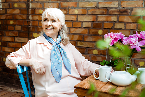 cheerful senior woman smiling while sitting near cups and teapot