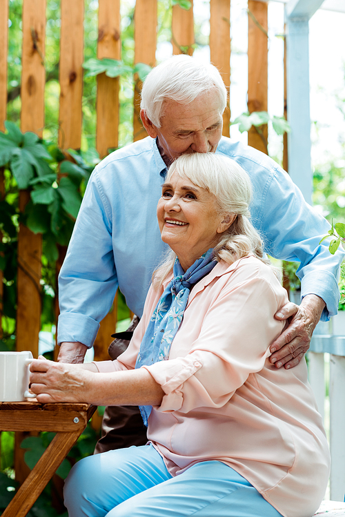 senior man with closed eyes kissing cheerful wife with grey hair