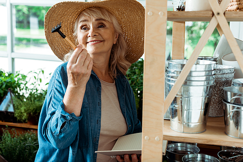 cheerful senior woman in straw hat holding small rake and notebook