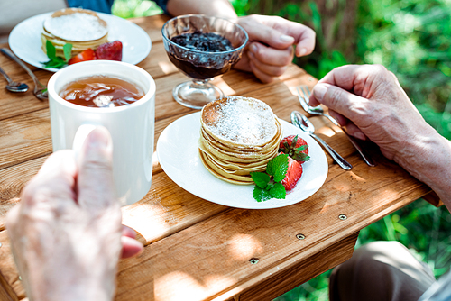 cropped view of senior man holding cup of tea near wife and tasty pancakes