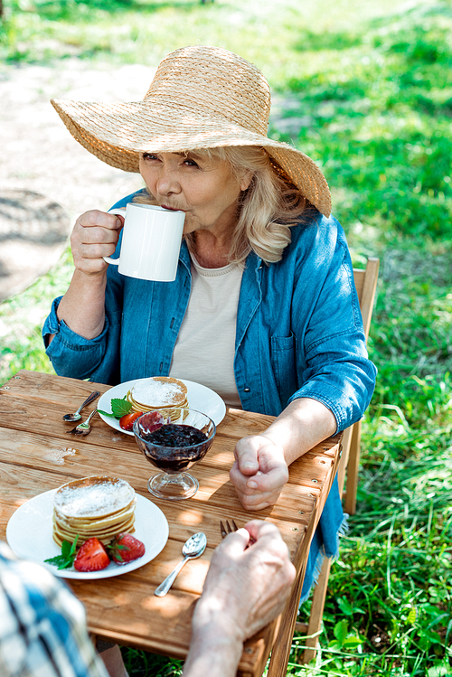 senior woman in straw hat drinking tea near sweet and tasty pancakes