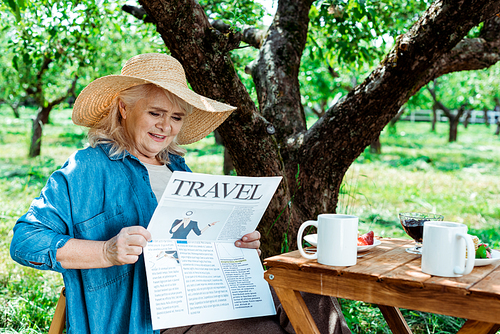 senior woman in straw hat sitting near tree and reading travel newspaper