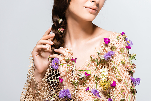 partial view of beautiful woman touching braid in mesh with spring wildflowers isolated on grey