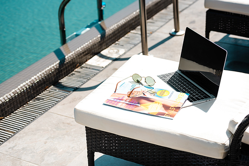 laptop with blank screen, magazine and sunglasses on sun bed near swimming pool