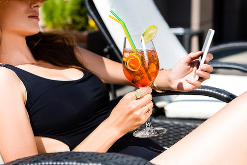 cropped view of girl in swimming suit with cocktail on sunbed using smartphone