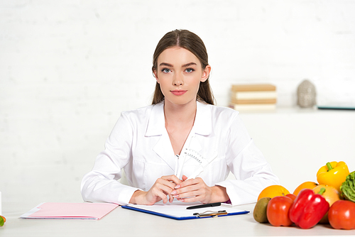 front view of dietitian in white coat at workplace with vegetables, folder and clipboard on table