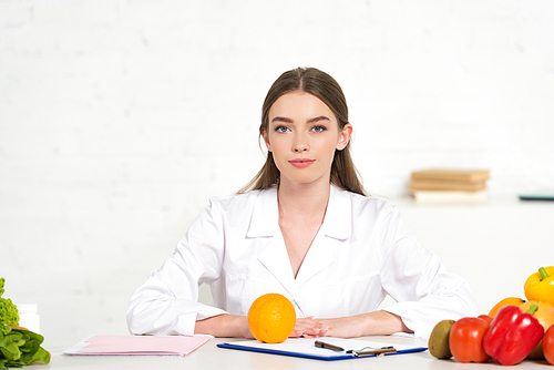 front view of dietitian in white coat at workplace with vegetables, folder and clipboard on table