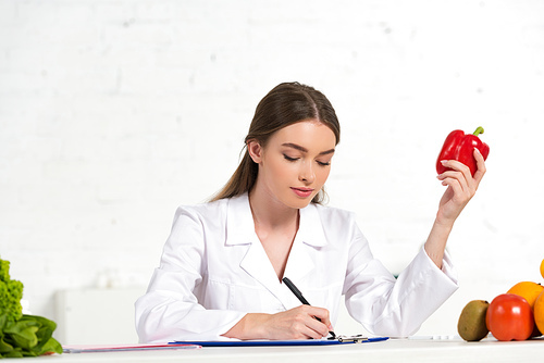 dietitian in white coat holding red bell pepper and writing in clipboard at workplace