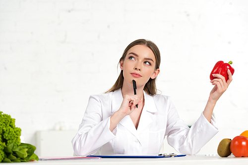 pensive dietitian in white coat holding red bell pepper and pen and looking up at workplace