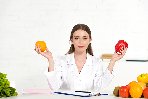 front view of dietitian in white coat holding orange and bell pepper at workplace