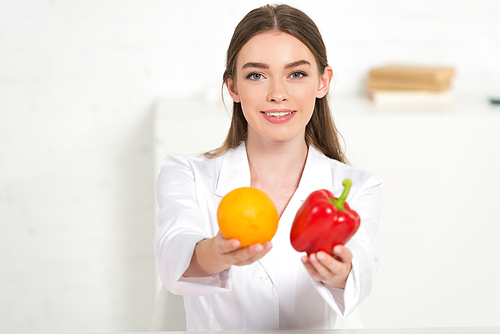 front view of smiling dietitian in white coat holding orange and red bell pepper