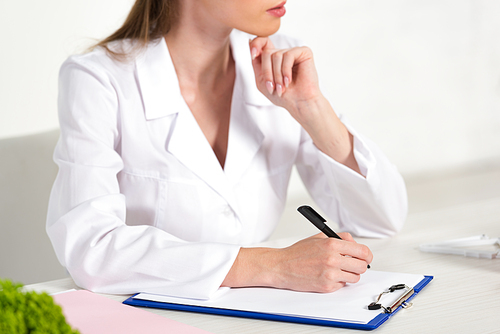 partial view of dietitian in white coat writing in clipboard at workplace