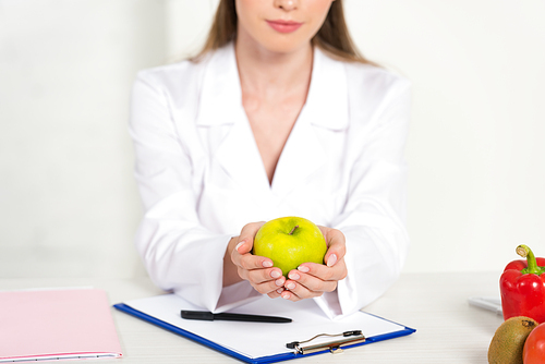 partial view of dietitian in white coat holding apple at workplace
