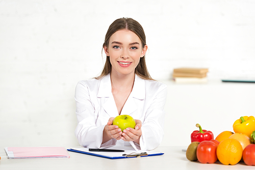 front view of smiling dietitian in white coat holding apple at workplace
