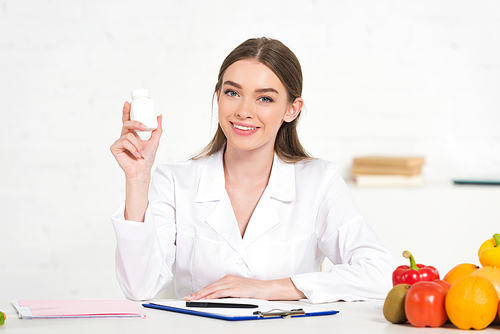 smiling dietitian in white coat holding pills at workplace with fruits and vegetables on table