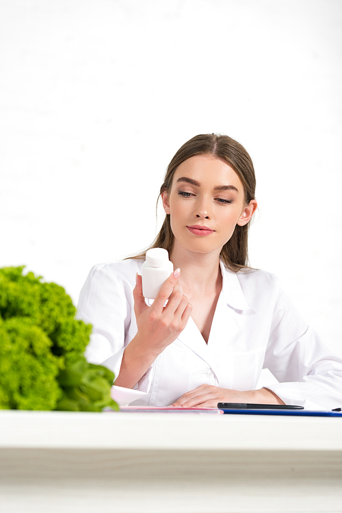 pensive dietitian in white coat holding pills at workplace