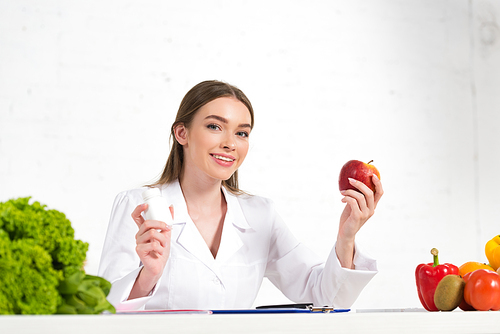 smiling dietitian in white coat holding pills and apple at workplace