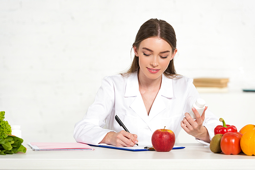 smiling dietitian in white coat holding pills and writing in clipboard at workplace with fruits and vegetables on table