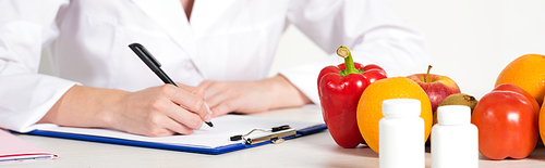 panoramic shot of dietitian in white coat writing in clipboard at workplace with pills, fruits and vegetables on table