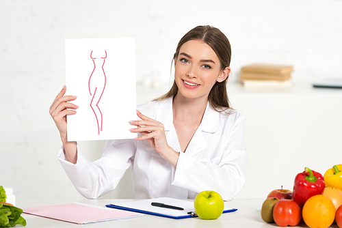 smiling dietitian in white coat holding paper with image of perfect body at workplace with fruits and vegetables on table