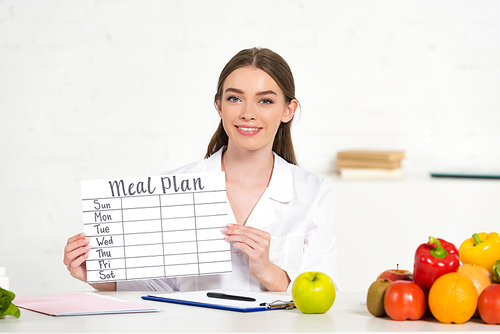 front view of smiling dietitian in white coat holding meal plan at workplace with fruits and vegetables on table