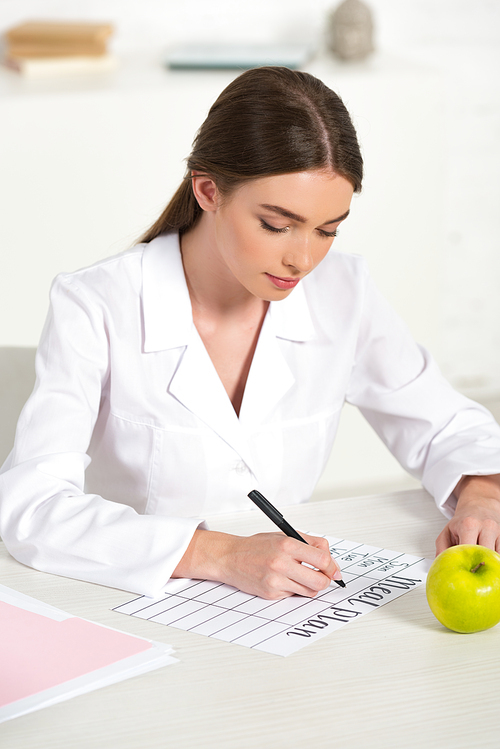 focused dietitian in white coat writing meal plan at table with green apple