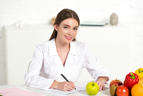 smiling dietitian in white coat writing at workplace with fruits and vegetables on table