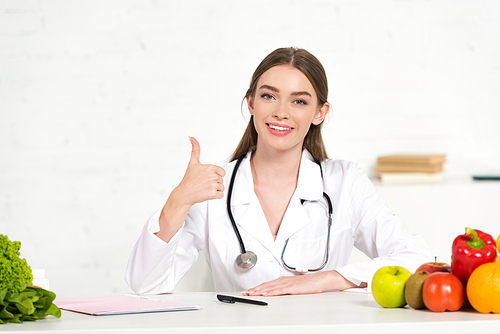 front view of smiling dietitian in white coat with stethoscope showing thumb up at workplace with fruits and vegetables on table