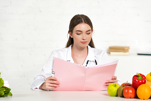 focused dietitian in white coat reading folder at workplace with fruits and vegetables on table