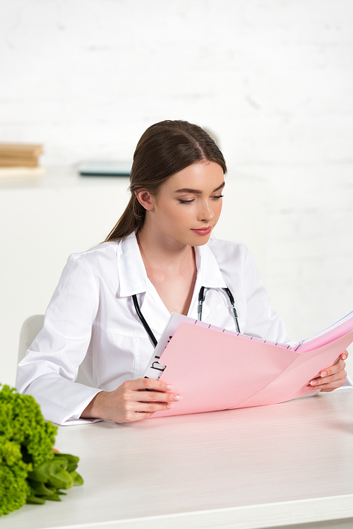 focused dietitian in white coat reading folder at workplace