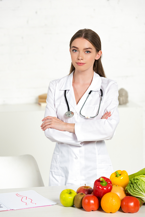 attractive dietitian in white coat with equipment standing with crossed arms near table with fresh fruits and vegetables