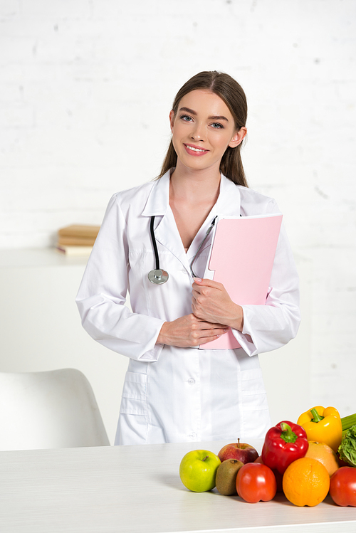 smiling attractive dietitian in white coat with equipment holding folder near table with fresh fruits and vegetables