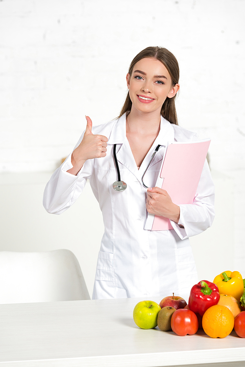 smiling dietitian in white coat holding folder and showing thumb up near table with fresh fruits and vegetables