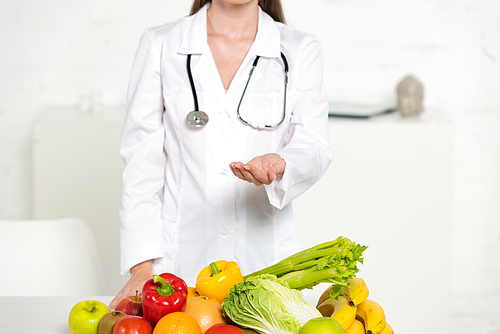 cropped view of dietitian in white coat with stethoscope standing with outstretched hand near fresh fruits and vegetables