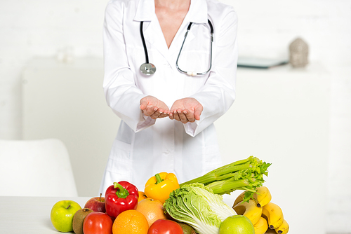 partial view of dietitian in white coat with stethoscope near fresh fruits and vegetables