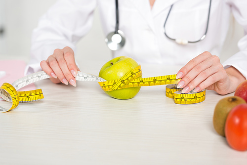cropped view of dietitian in white coat holding measure tape at table with fruits