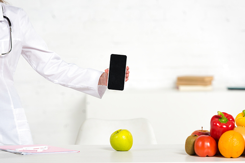 cropped view of dietitian in white coat holding smartphone with blank screen near table with fruits and vegetables